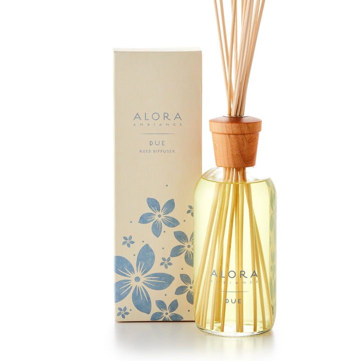 Alora Ambiance 16oz Due reed diffuser with glass bottle and wooden cap next to tan box with purple floral pattern on it.