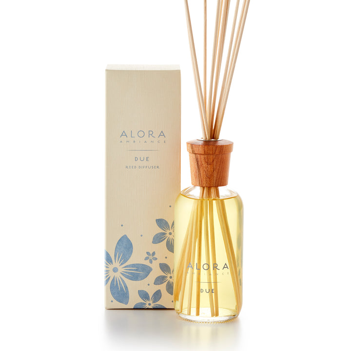 Alora Ambiance 8oz Due reed diffuser with glass bottle and wooden cap next to tan box with purple floral pattern on it.