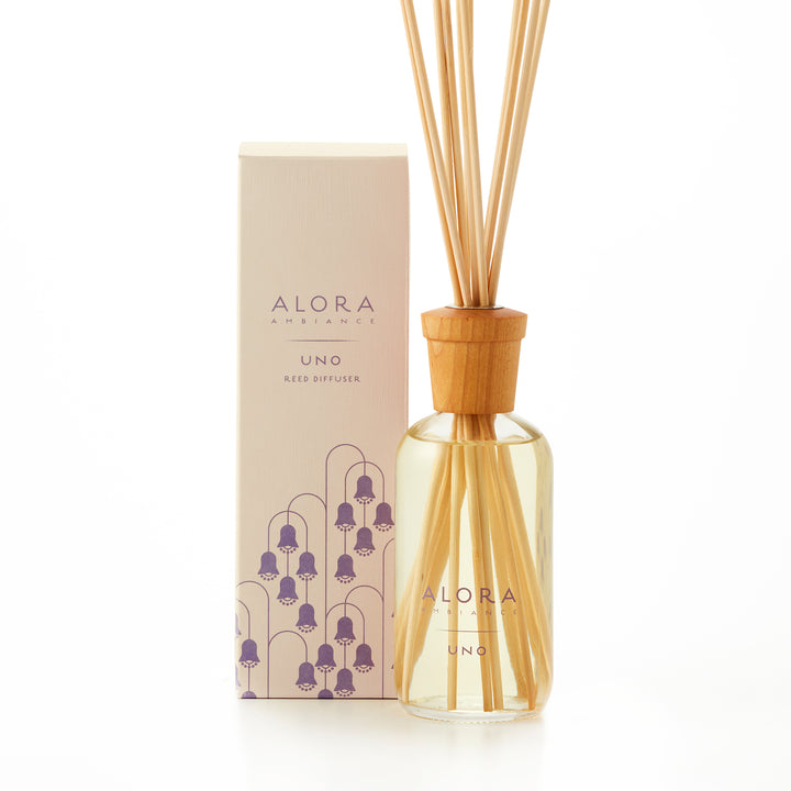 Alora Ambiance 8oz Uno reed diffuser with glass bottle and wooden cap next to tan box with purple Lily of the Valley flower pattern on it.