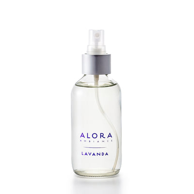 Small, glass spray bottle with "Alora Ambiance" and "Lavanda" written in purple font on the front, bottle is filled with clear, liquid fragrance