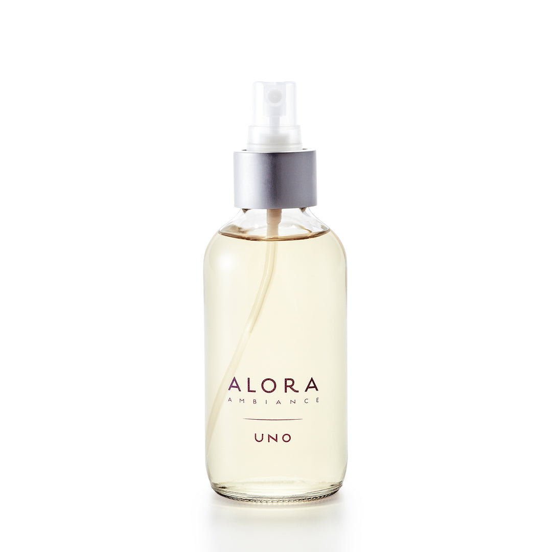 Small, glass spray bottle with the words "Alora Ambiance" and "Uno" written on the front in purple font, bottle is filled with very pale yellow liquid fragrance