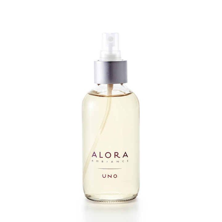 Small, glass spray bottle with the words "Alora Ambiance" and "Uno" written on the front in purple font, bottle is filled with very pale yellow liquid fragrance