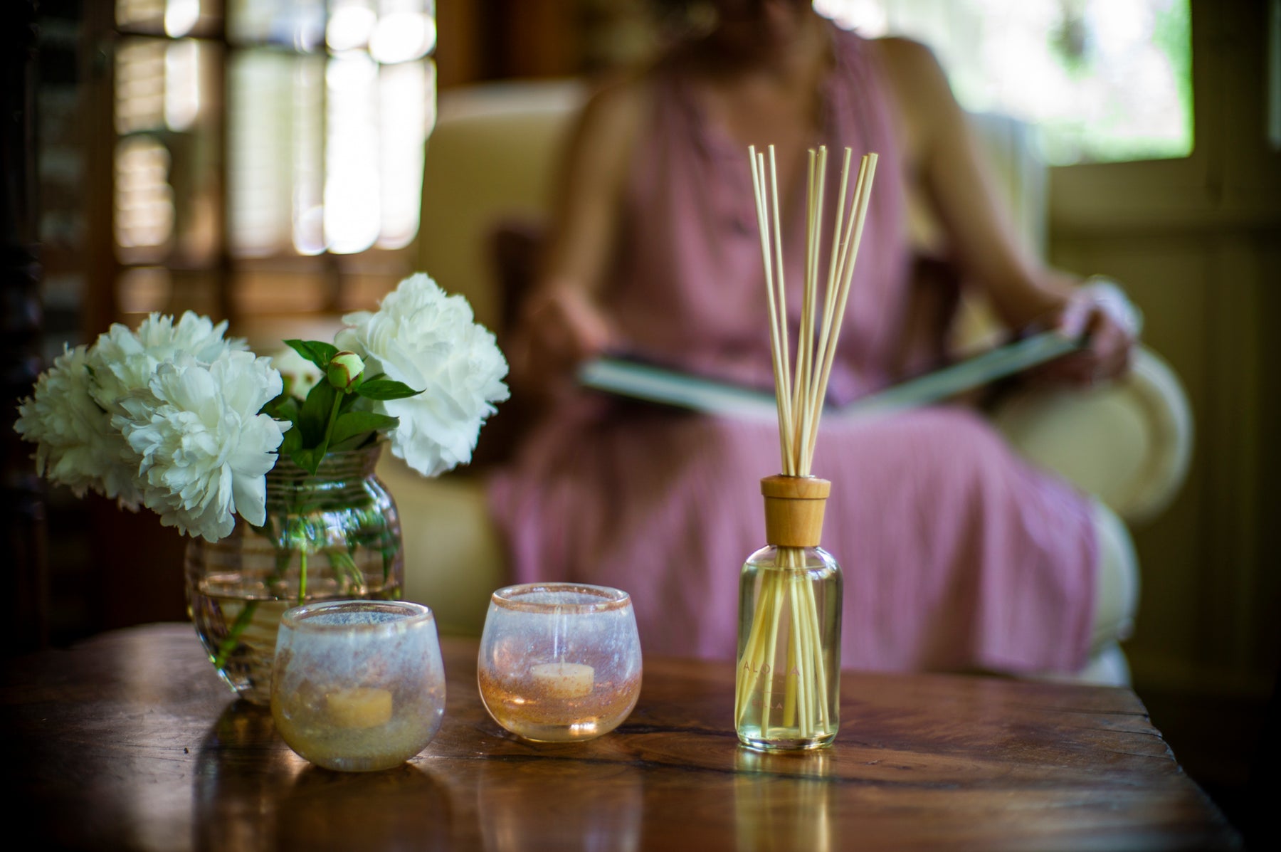 Woman sitting in pink dress reading by coffee table with a reed diffuser on the table.