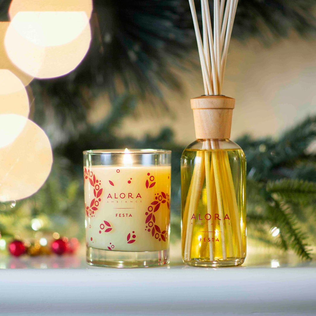 Festa candle and reed diffuser on mantle in front of pine greens and twinkle lights