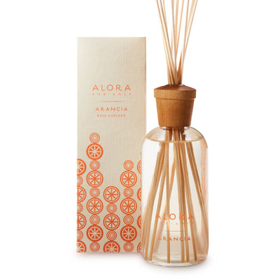 Glass bottle filled with clear, liquid fragrance, topped with wood cap with reeds passing through cap into liquid. Bottle next to tan box says “Alora Ambiance” and “Arancia”