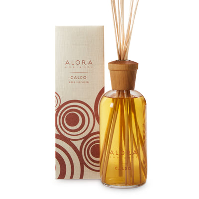 Glass bottle filled with gold, liquid fragrance, topped with wood cap with reeds passing through cap into liquid. Bottle says “Alora Ambiance” and “Caldo” and is by a tan box 