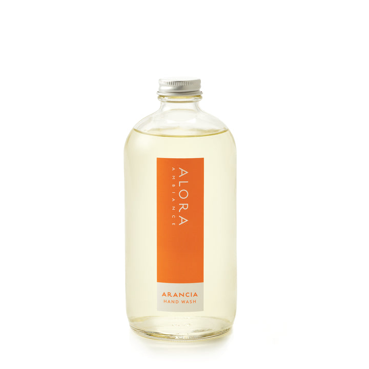 Tall glass bottle topped with a metal cap and filled with clear, liquid hand wash with label that says “Alora Ambiance” and “Arancia Hand Wash” on the front