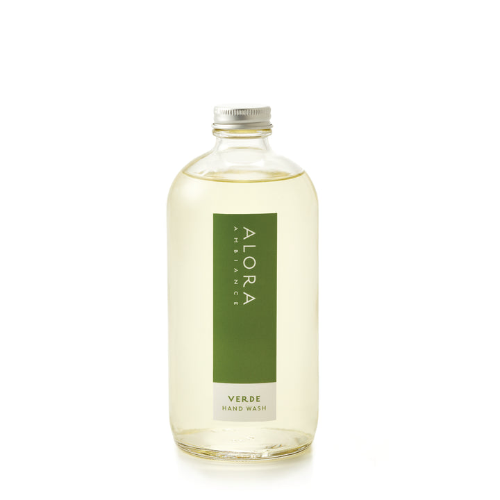 Tall glass bottle topped with a metal cap and filled with clear, liquid hand wash with label that says “Alora Ambiance” and “Verde Hand Wash” on the front