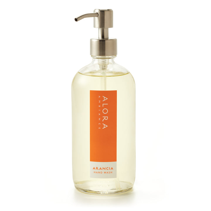 Tall glass bottle topped with a metal pump and filled with clear, liquid hand wash with label that says “Alora Ambiance” and “Arancia Hand Wash” on the front