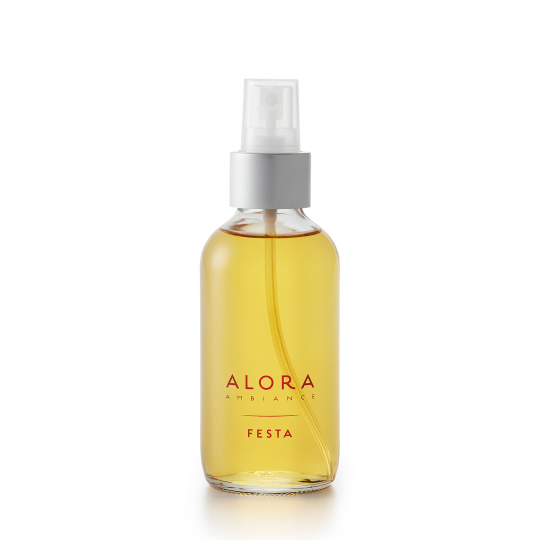 Small, glass spray bottle with "Alora Ambiance" and "Festa" written on the front in red font, bottle is filled with amber-colored liquid