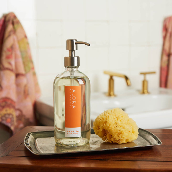 Arancia hand wash bottle on silver tray with sponge by bathroom sink.