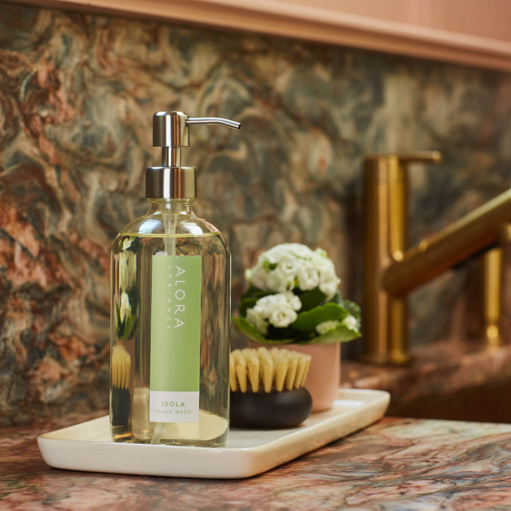 Isola hand wash bottle on a tray on a granite kitchen countertop next to sink. Tray has a scrub brush and small pot of flowers on it