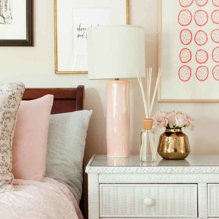 Rosa diffuser on nightstand next to a gold vase filled with roses, a pink lamp, and a bed with pink sheets
