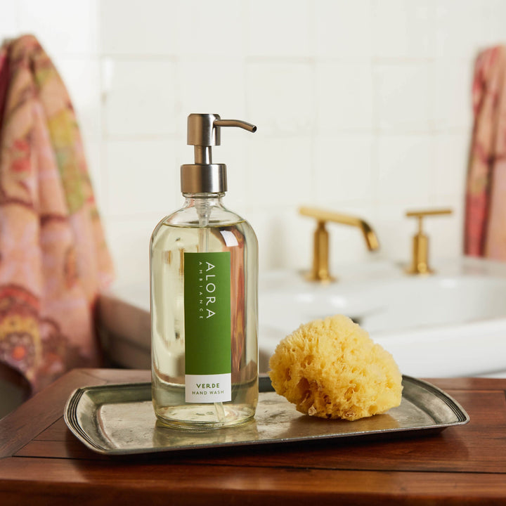 Verde hand wash bottle sites on a silver tray next to a sponge. A bathroom sink and two pink towels are in the background.