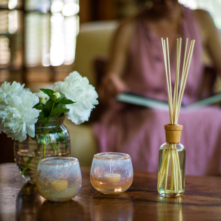 Bella reed diffuser on table next to two candle votives, a vase of white flowers, with women in purple dress reading in the background