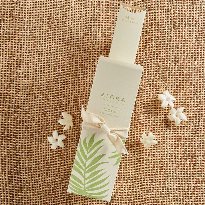 Tall tan box that says "Alora Ambiance" and "Isola Reed Diffuser" is wrapped in a tan ribbon and sitting on a packet of reeds. Five small white flowers sit nearby.