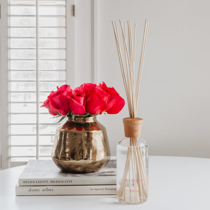 Rosa reed diffusers, next to gold vase filled with pink roses, stacked on two books