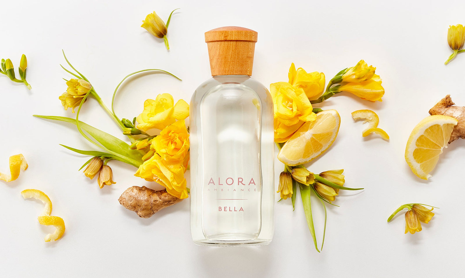 Bella diffuser bottle laying next to ylang ylang flowers, lemon slices, and ginger pieces