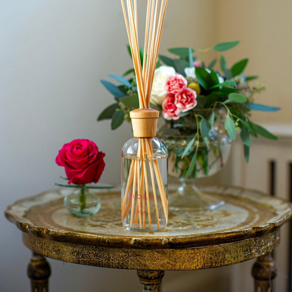Rosa diffuser on a table next to a bouquet of greens and roses and a bud vase with a pink rose in it