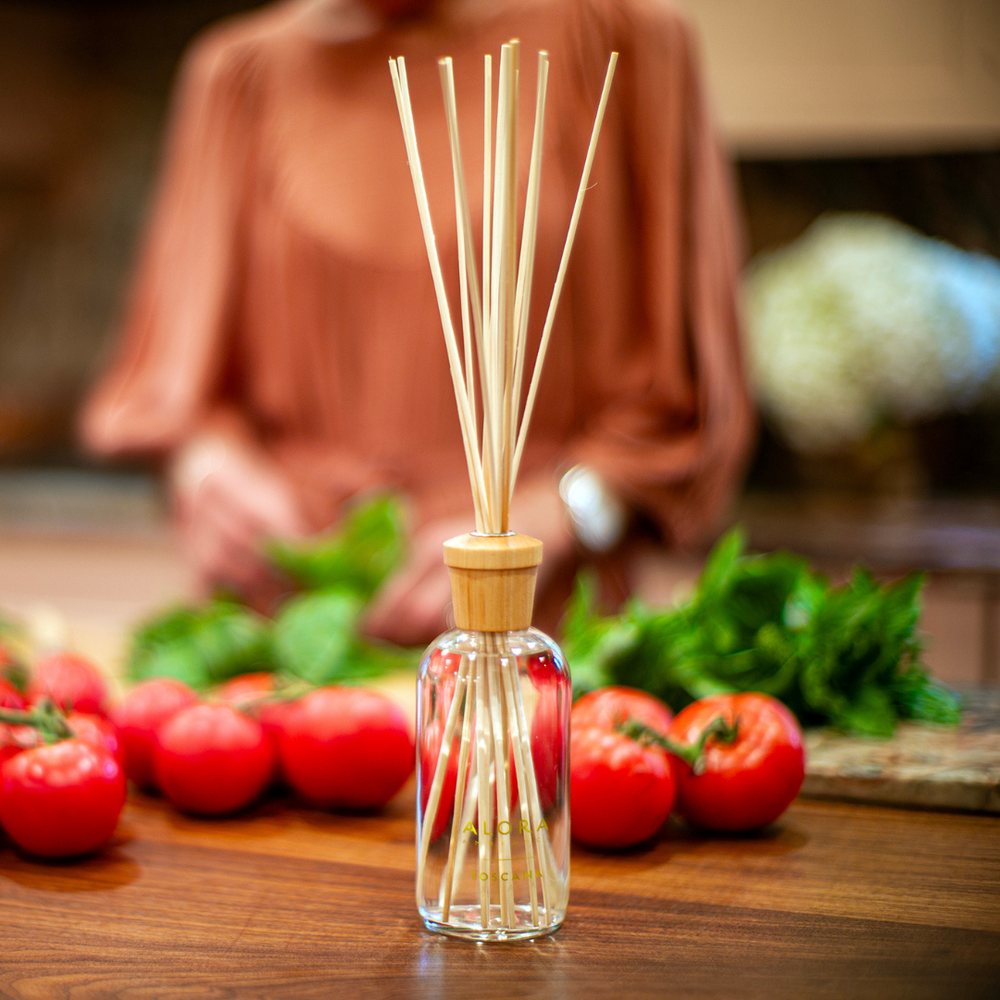 Toscana reed diffuser on butcher block countertop in front of tomatoes and woman sorting basil