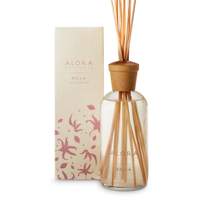 Glass bottle filled with clear, liquid fragrance, topped with wood cap with reeds passing through cap into liquid. Bottle next to tan box says “Alora Ambiance” and “Bella”