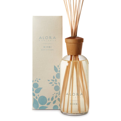 Glass bottle filled with clear, liquid fragrance, topped with wood cap with reeds passing through cap into liquid. Bottle next to tan box says “Alora Ambiance” and “Bimbi”