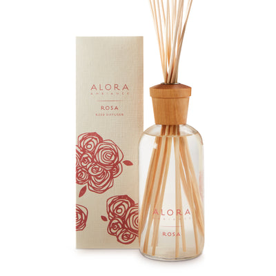 Glass bottle filled with liquid fragrance, topped with wood cap with reeds passing through cap into liquid. Bottle says “Alora Ambiance” and “Rosa” and is next to tan box 