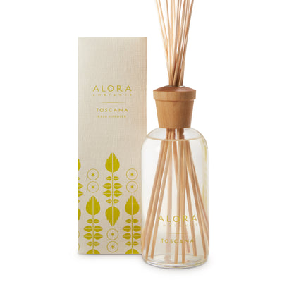 Glass bottle filled with clear, liquid fragrance, topped with wood cap with reeds passing through cap into liquid. Bottle next to tan box says “Alora Ambiance” and “Toscana”