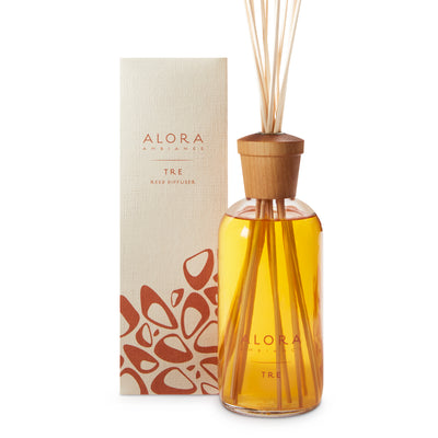 Glass bottle filled with golden, liquid fragrance, topped with wood cap with reeds passing through cap into liquid. Bottle says “Alora Ambiance” and “Tre” and is next to tan box 