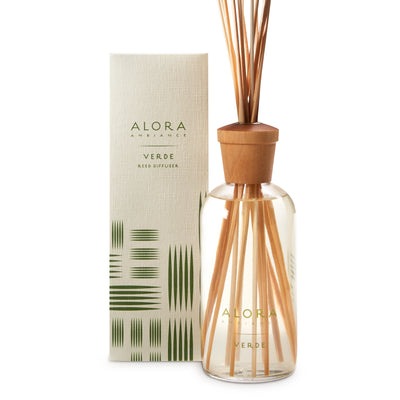 Glass bottle filled with clear, liquid fragrance, topped with wood cap with reeds passing through cap into liquid. Bottle next to tan box says “Alora Ambiance” and “Verde”