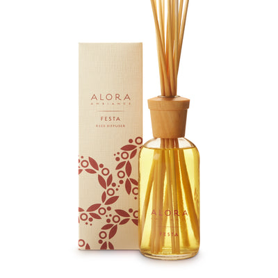 Glass bottle filled with golden, liquid fragrance, topped with wood cap with reeds passing through cap into liquid. Bottle says “Alora Ambiance” and “Festa” and is next to tan box
