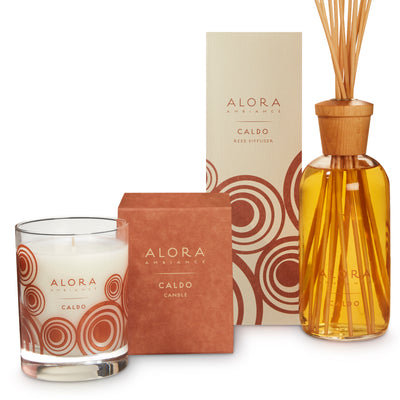 Candle in glass jar that says “Alora Ambiance” and “Caldo,” next to a rust-colored square box by tall tan box that says “Alora Ambiance” and “Caldo Reed Diffuser” by reed diffuser