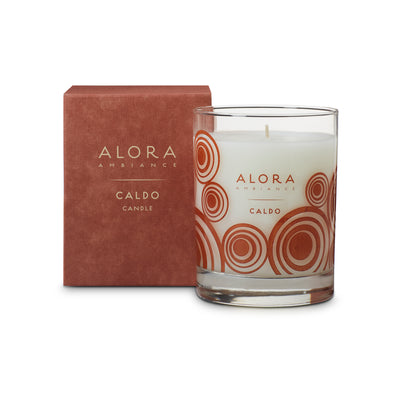 White-colored candle in glass jar that says “Alora Ambiance” and “Caldo” and is next to rust colored box that says “Alora Ambiabnce” and “Caldo candle”