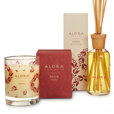 Candle in glass jar that says “Alora Ambiance” and “Festa,” next to a red square box next to a tall tan box that says “Alora Ambiance” and “Festa Reed Diffuser” by glass reed diffuser