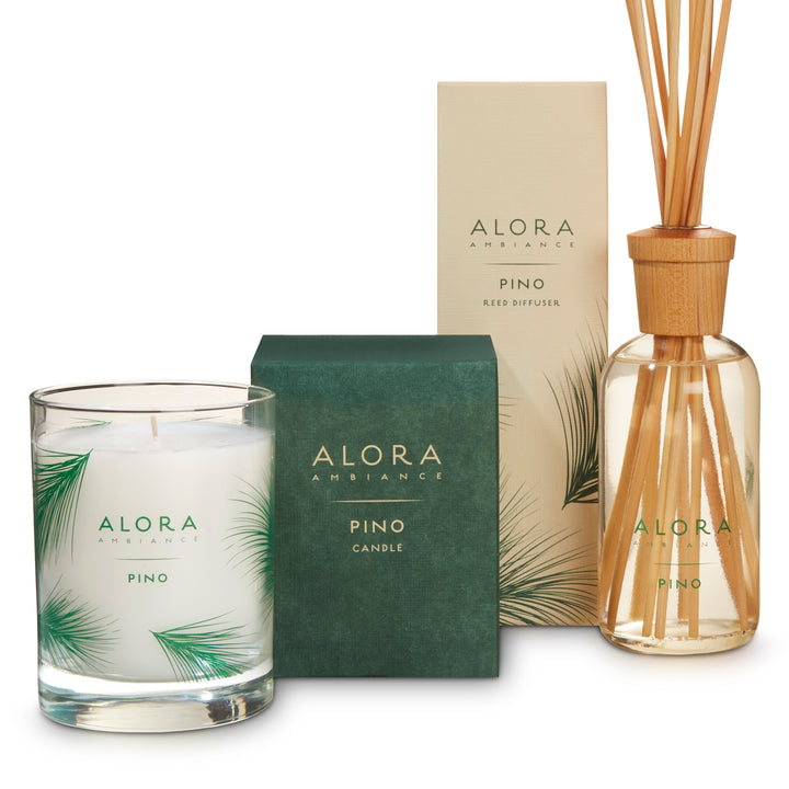 Candle in glass jar that says “Alora Ambiance” and “Pino,” next to a green square box next to a tall tan box that says “Alora Ambiance” and “Pino Reed Diffuser” by reed diffuser