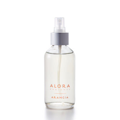 Small, glass spray bottle with "Alora Ambiance" and "Arancia" written on the front in orange font, bottle is filled with clear liquid fragrance