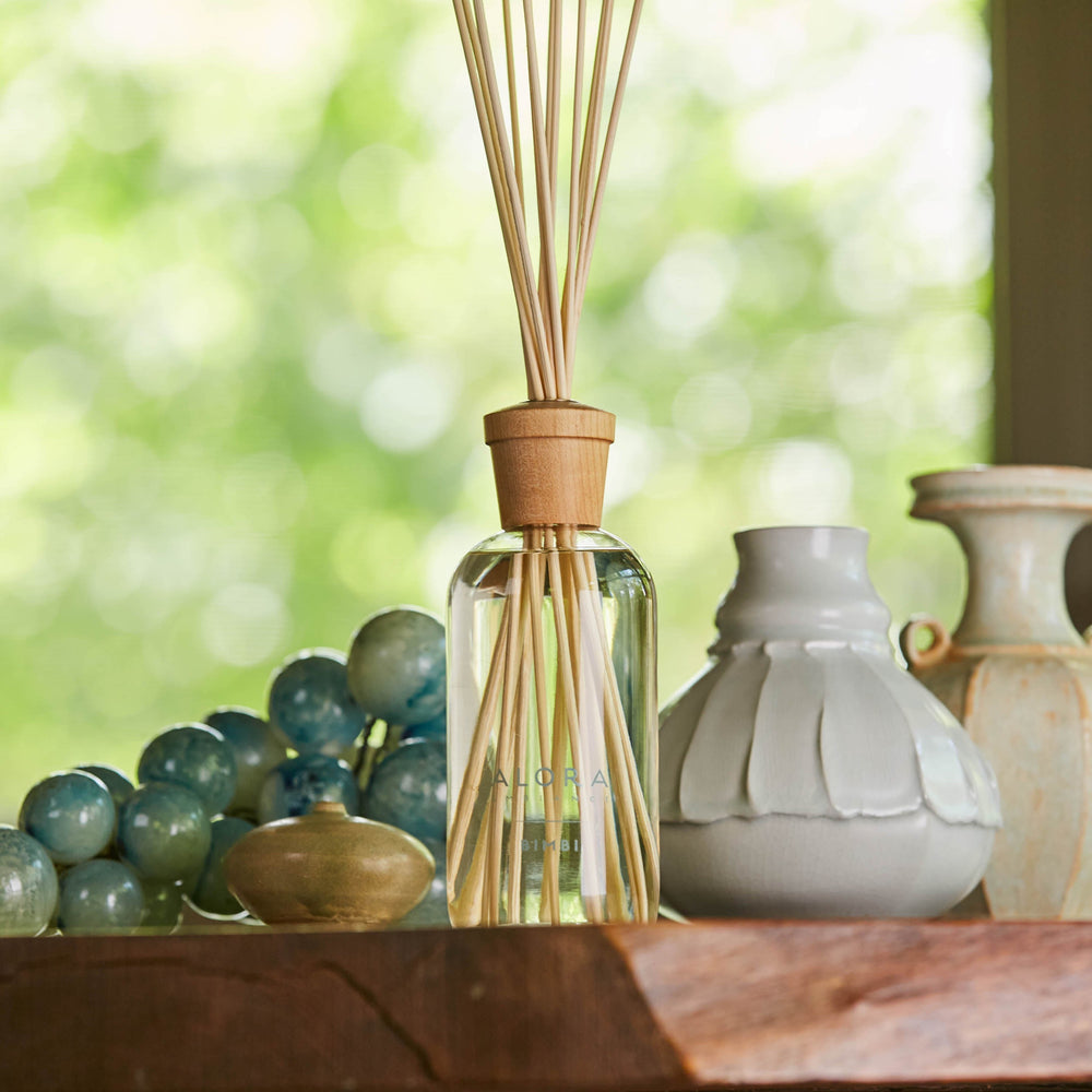 Bimbi reed diffuser on a wooden shelf alongside two white vases and a blue glass decor piece