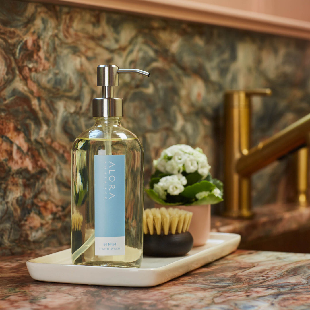 Bimbi hand wash bottle on a white porcelain tray next to a small, brown scrub brush and small pot of white flowers. Tray sits on a granite countertop next to a sink.