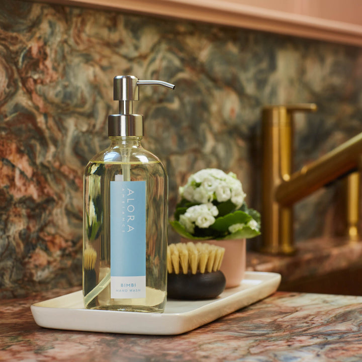 Bimbi hand wash bottle on a white porcelain tray next to a small, brown scrub brush and small pot of white flowers. Tray sits on a granite countertop next to a sink.