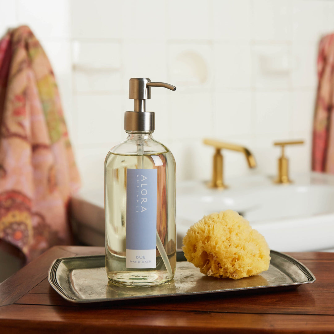 Due hand wash bottle sites on a silver tray next to a sponge. A bathroom sink and two pink towels are in the background.