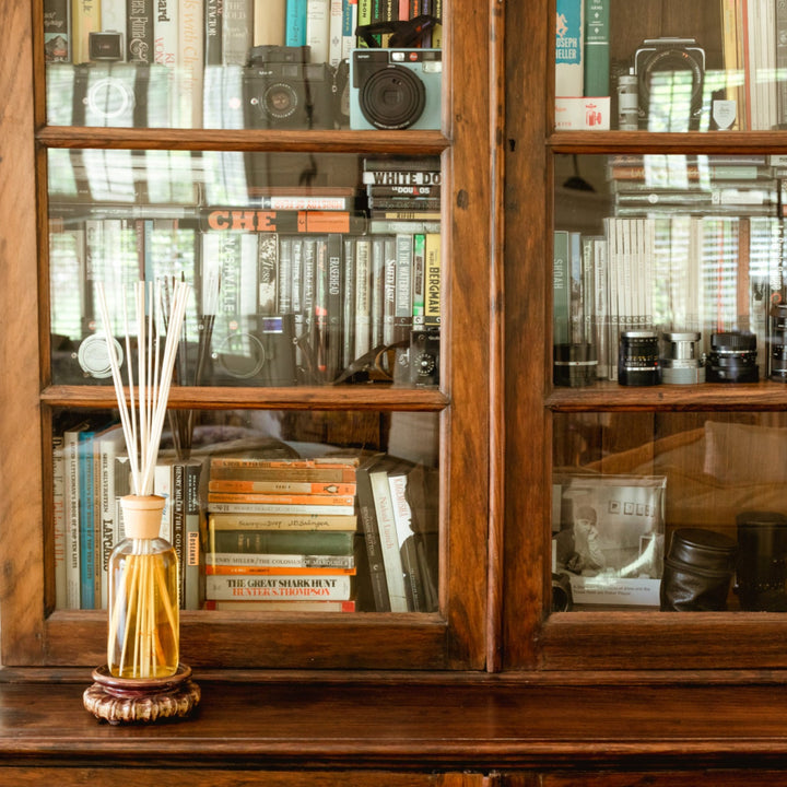 Caldo reed diffuser in front of wooden cupboard with glass doors. Shelves in cupboard are filled with books, old cameras, and knick knacks.