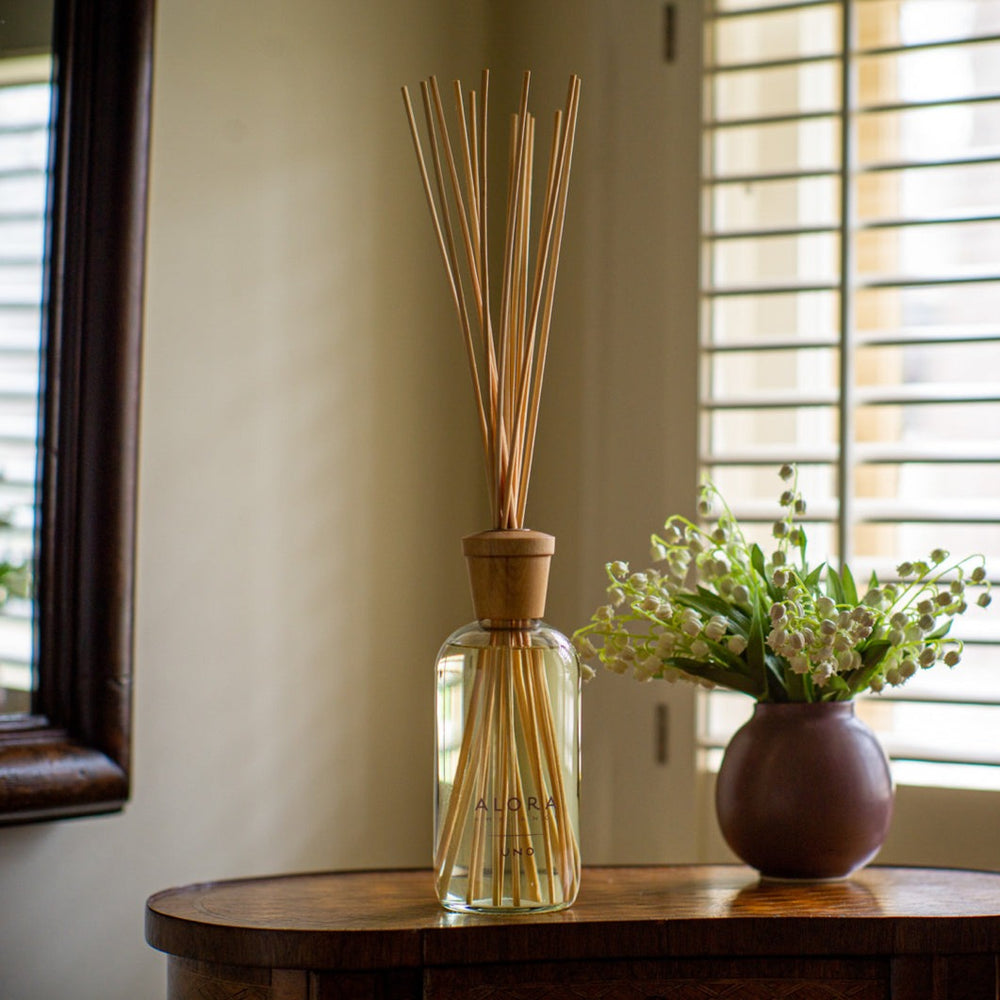 Uno reed diffuser on a small table next to a purple vase filled with lily of the valley flowers