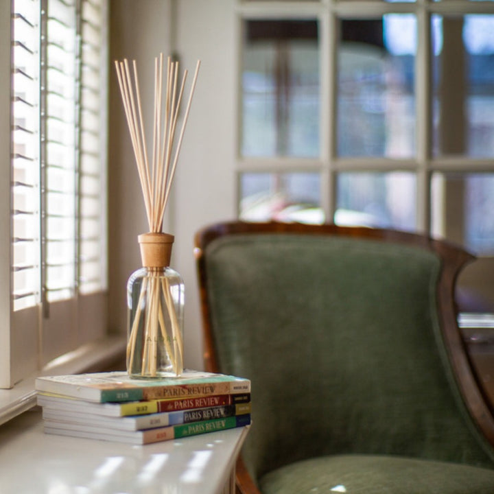 Verde reed diffuser on a stack of books next to a window and a green velvet covered chair