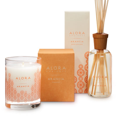 Candle in glass jar that says “Alora Ambiance” and “Arancia,” next to a orange square box by tall tan box that says “Alora Ambiance” and “Arancia Reed Diffuser” by reed diffuser